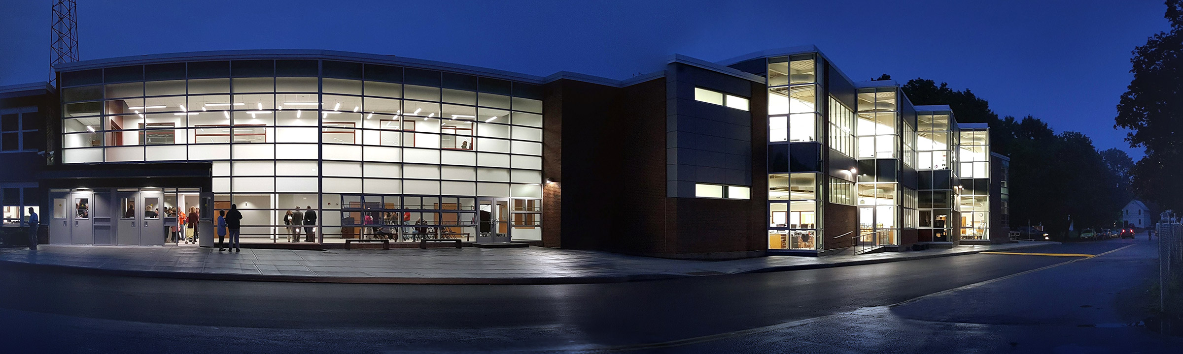 Panoramic photo of Burnt Hills-Ballston Lake High School building in the evening with brightly-lit windows and people