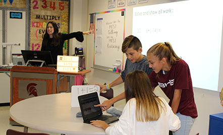 three students looking at a computer and teacher in background standing behind a smarthub pointing to a whiteboard