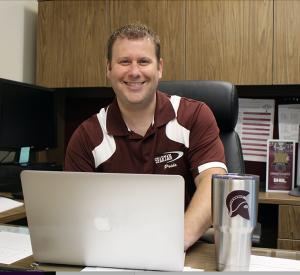 assistant superintendent wearing a maroon shirt sitting in a chair with a laptop on a desk