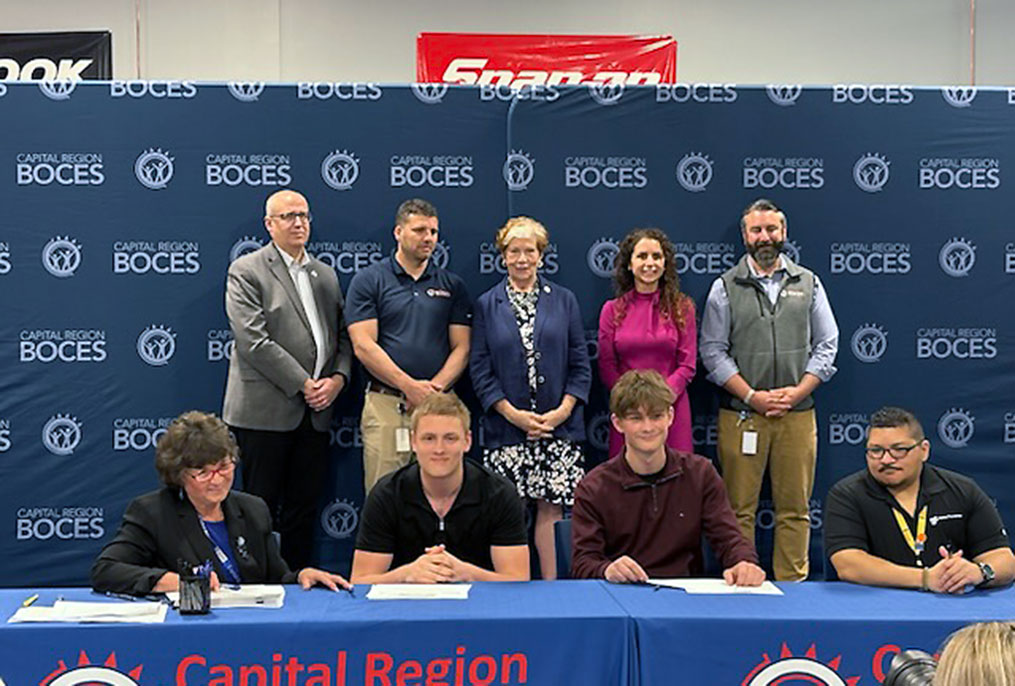 two students seated at a table signing papers with adults on either side and five adults standing behind the students' chairs