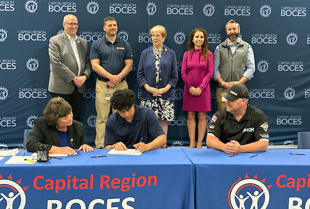 one student seated at a table and bent over signing a piece of paper with an adult on either side watching and five adults standing behind the table watching