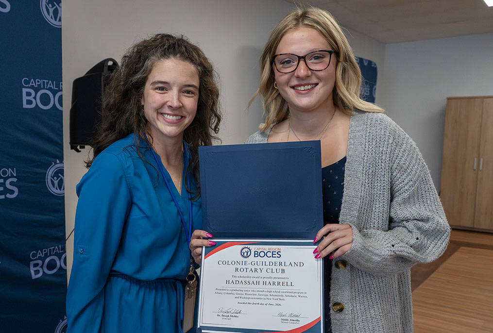 an adult in a blue dress standing with a student in a grey sweater who is holding a certificate
