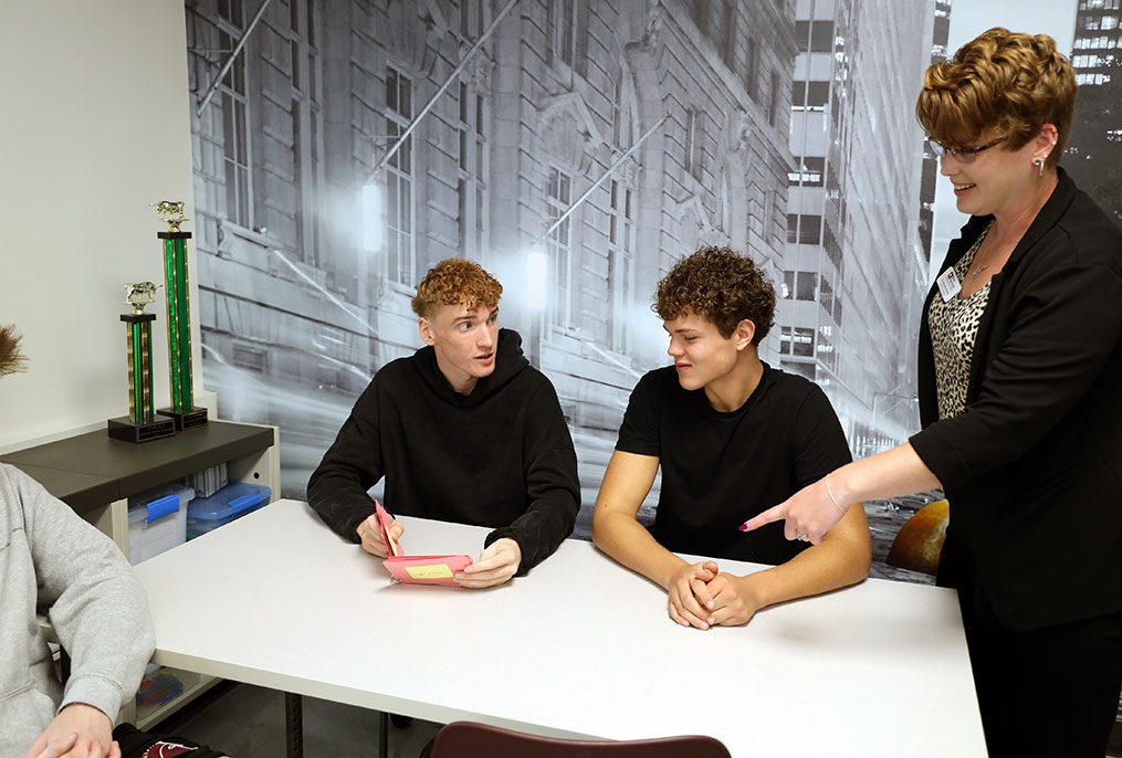 two students seated at a table and an adult is pointing to the index cards in one student's hands